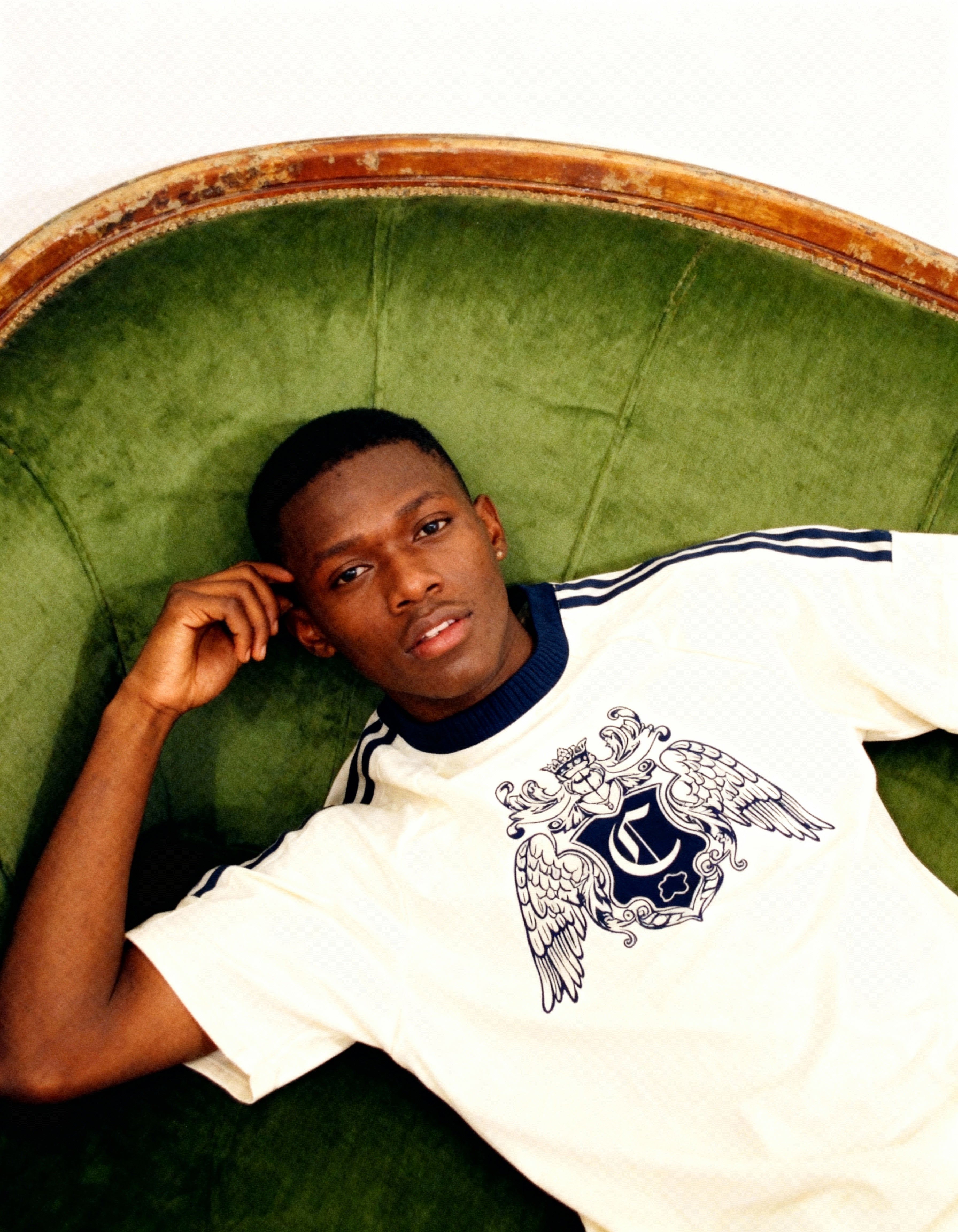 Jeune homme allongé sur un canapé vert, vêtu d’un t-shirt blanc à blason bleu, regardant calmement l’appareil photo.