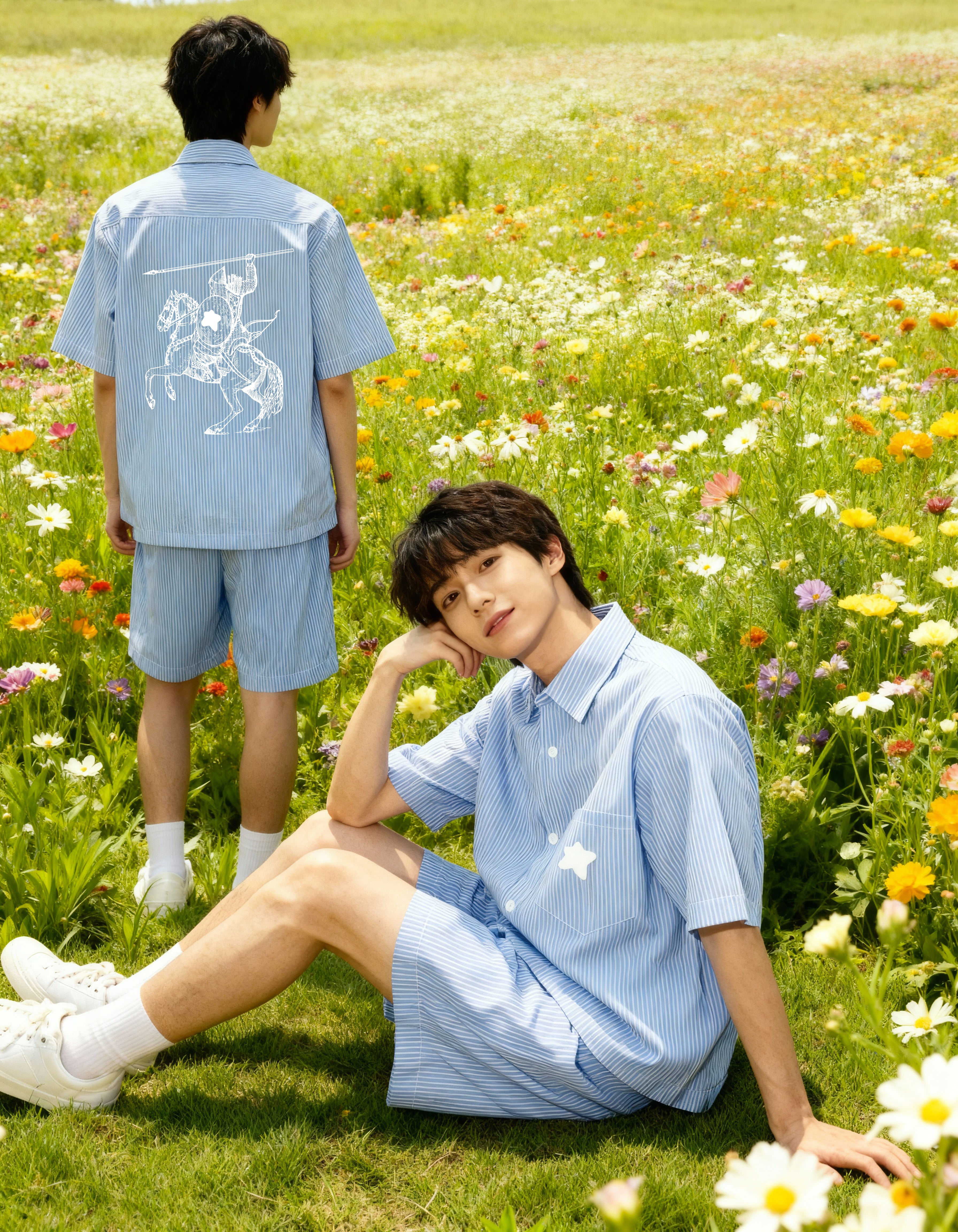 Deux jeunes hommes dans un champ fleuri, portant des ensembles à rayures bleues ; l’un assis souriant vers l’objectif, l’autre debout de dos avec une illustration de cavalier sur sa chemise.